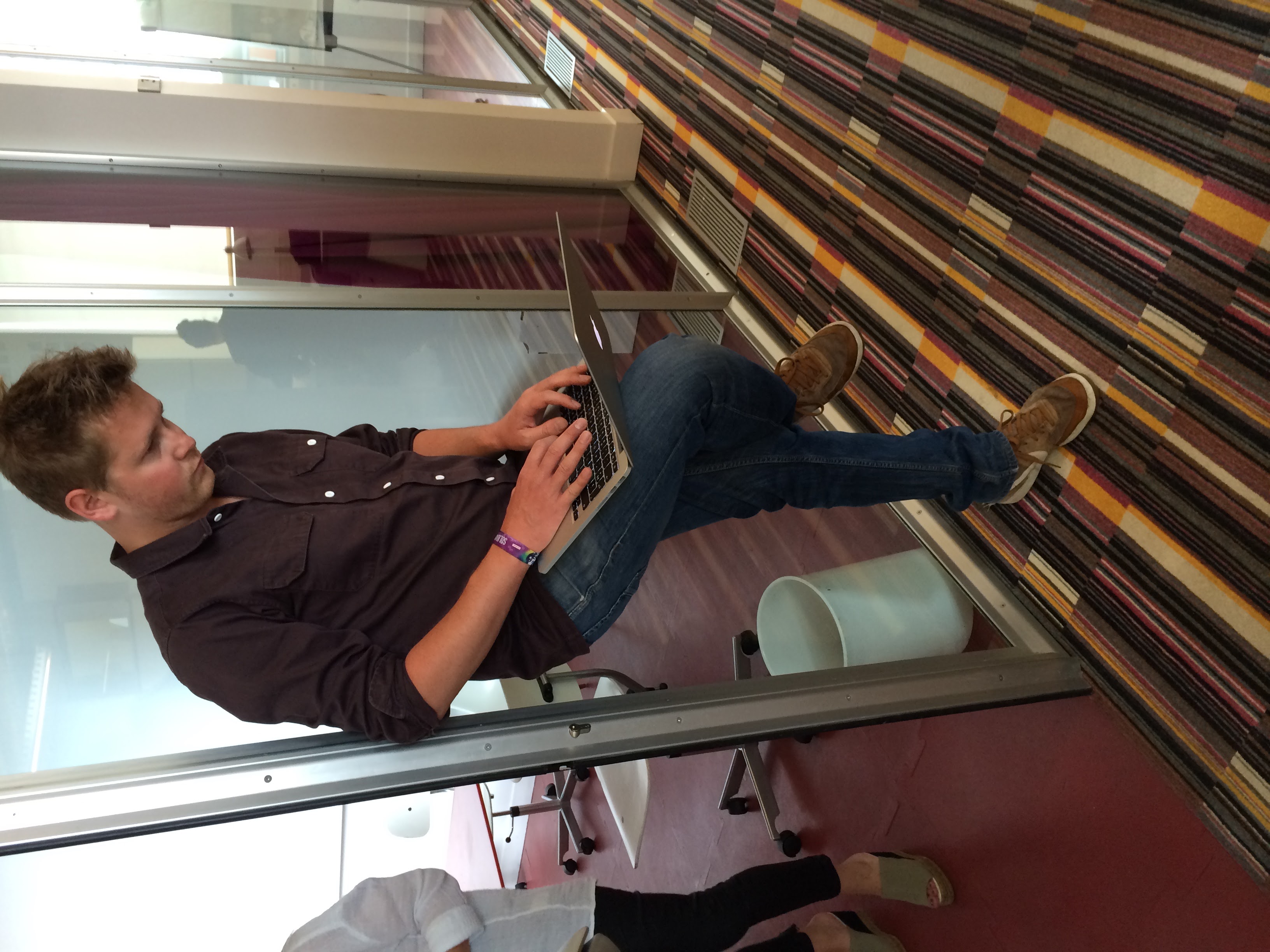 Maxime debout, posé sur un vitre d'une salle de réunion, avec l'ordi sur les genoux, en train de travailler dans une position improbable.