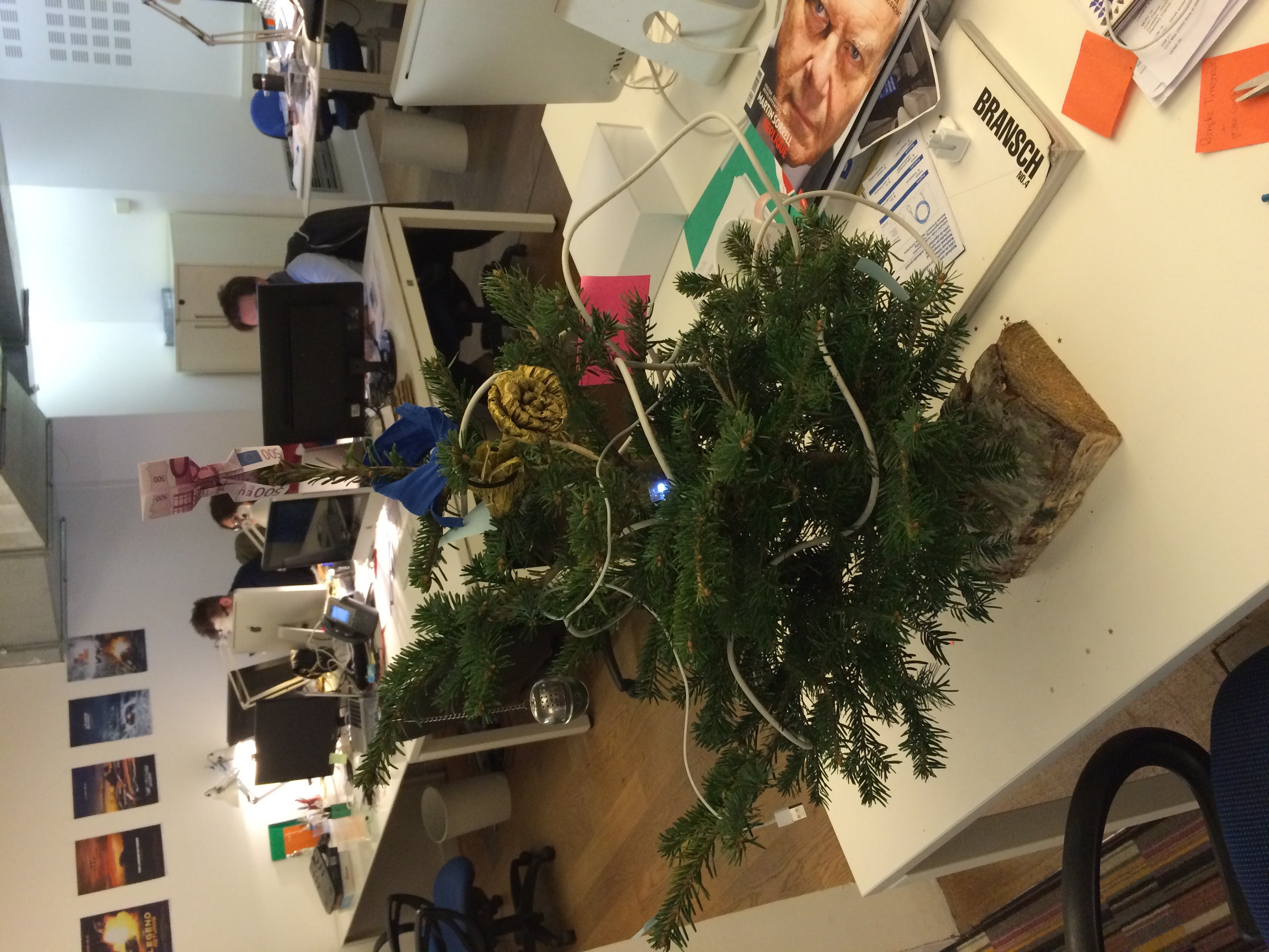 En premier plan, un sapin décoré avec des cables USB et Ethernet, dans un bureau avec des peronnes qui travaillent à l'ordinateur.