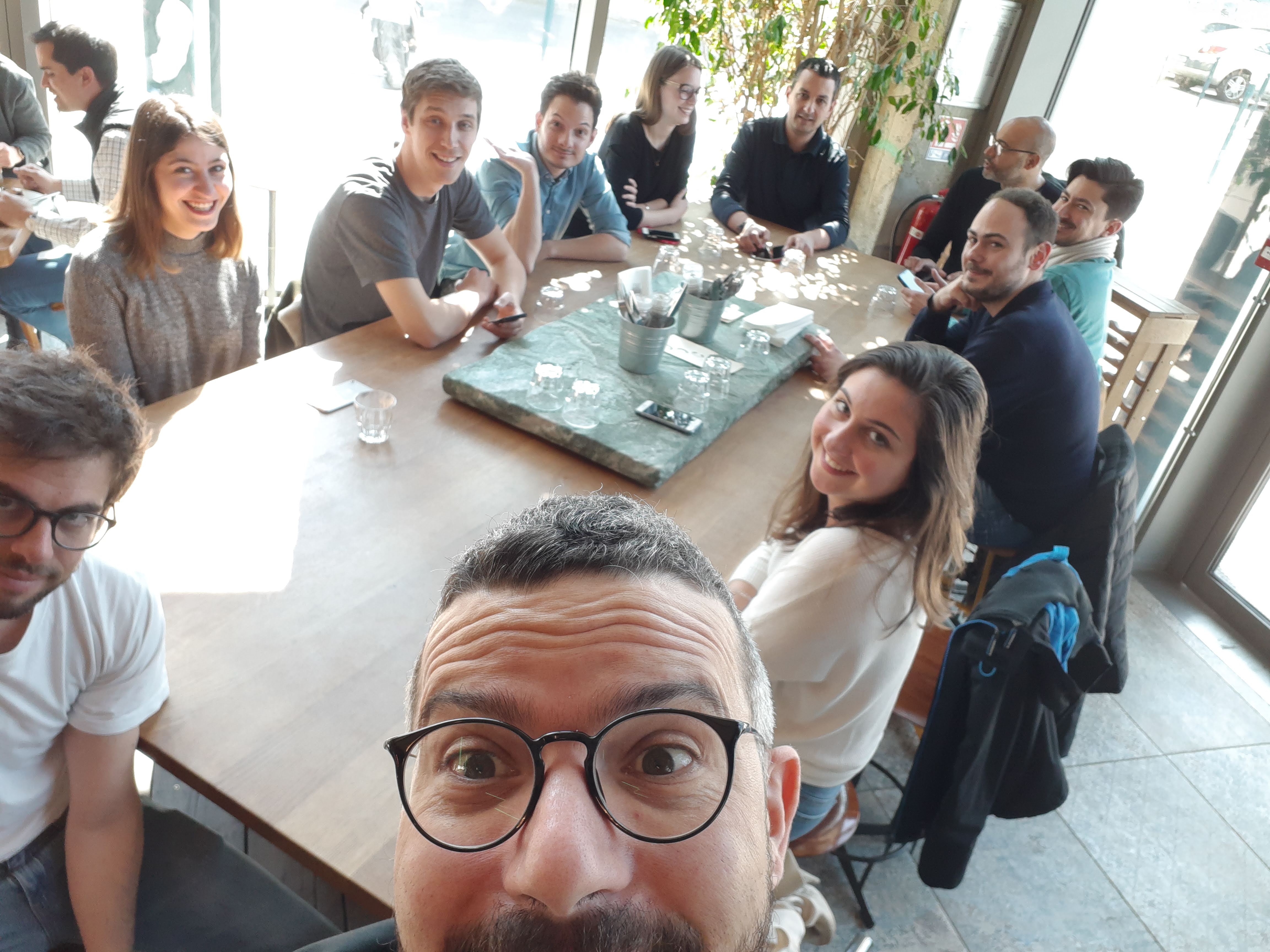 Équipe attablée dans un café lumineux, souriant à l’appareil photo lors d’un déjeuner.