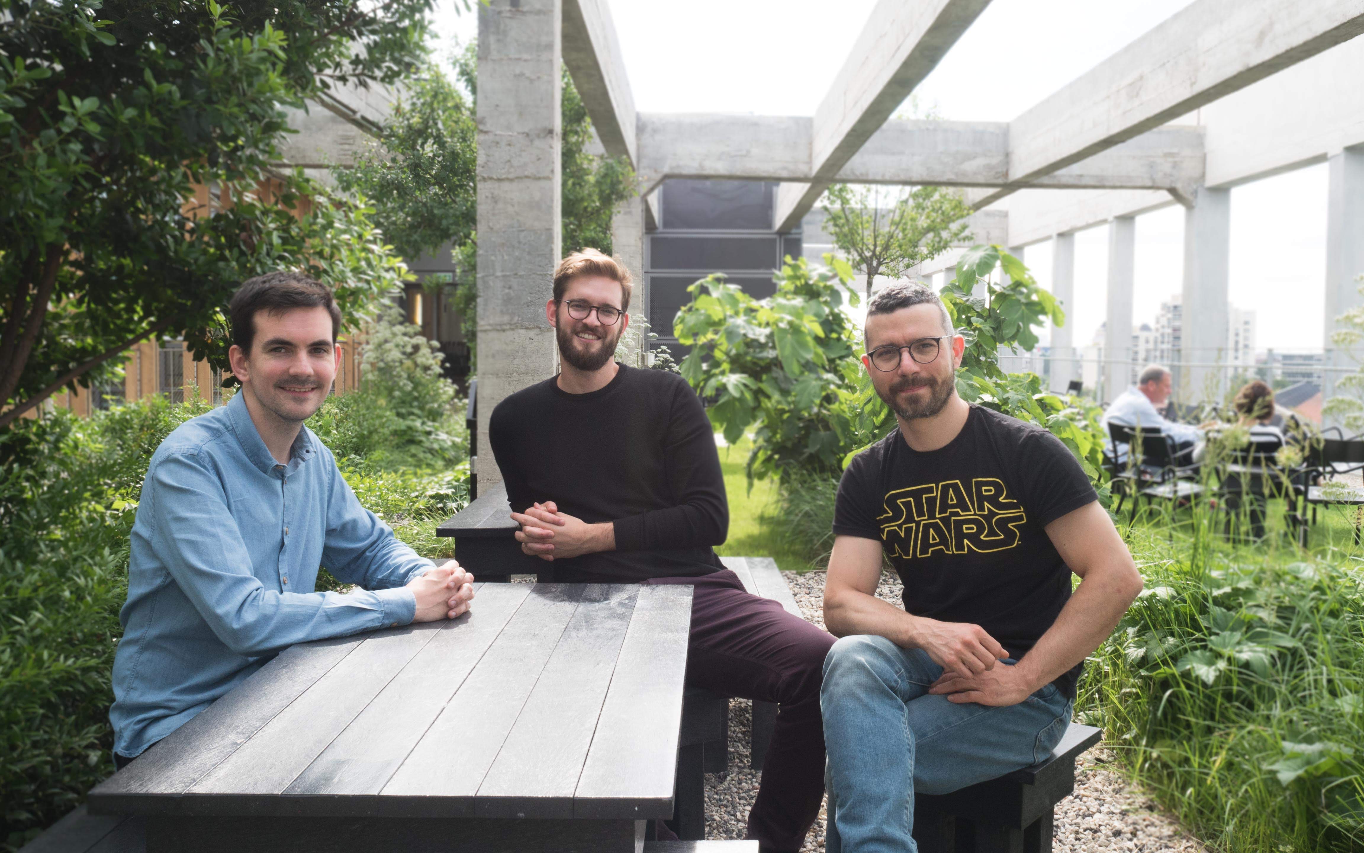 Trois hommes assis à une table extérieure entourée de verdure sur une terrasse moderne.