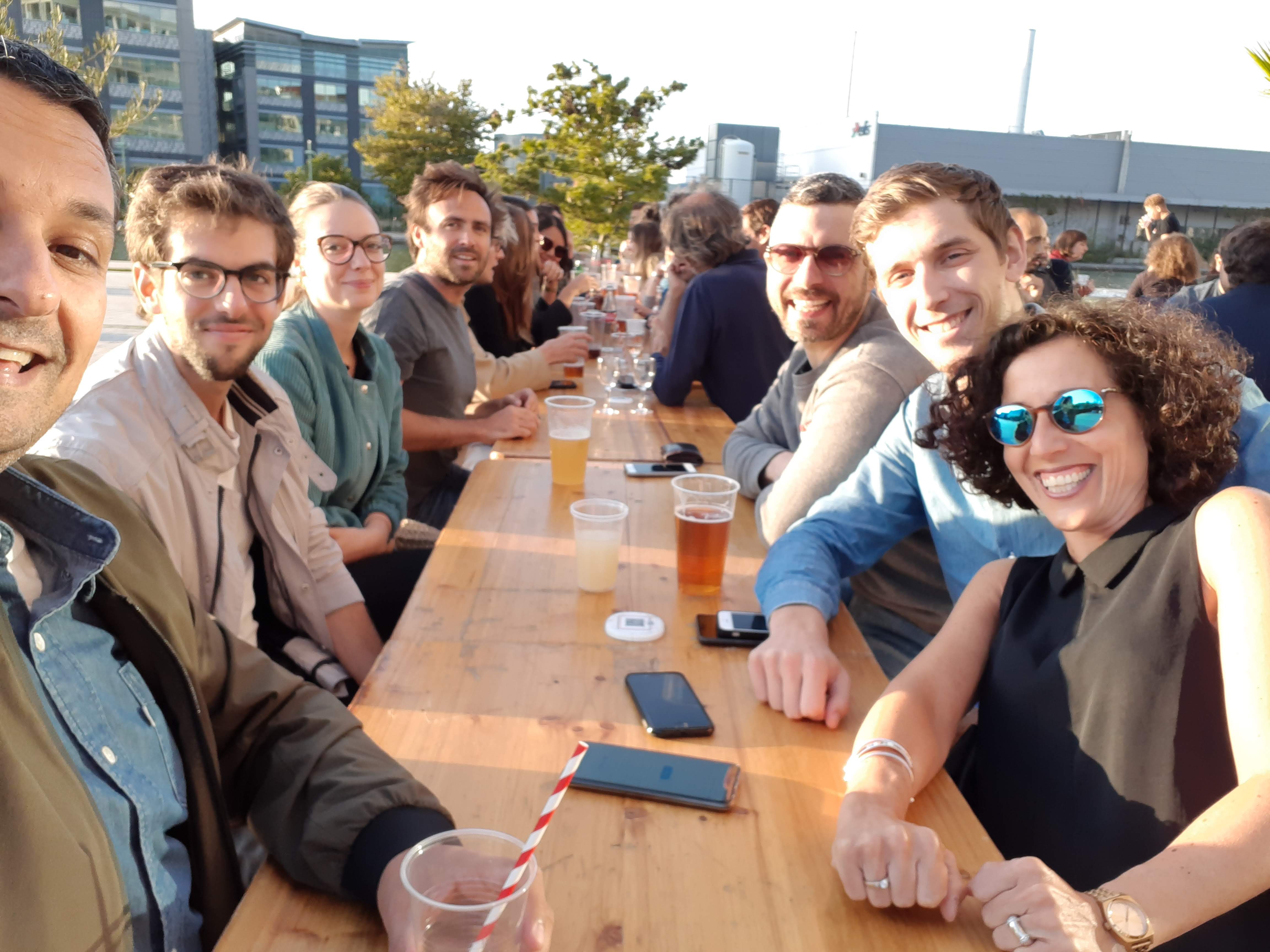 Groupe de collègues attablés en terrasse extérieure, verres et smartphones posés sur la table, ambiance ensoleillée.