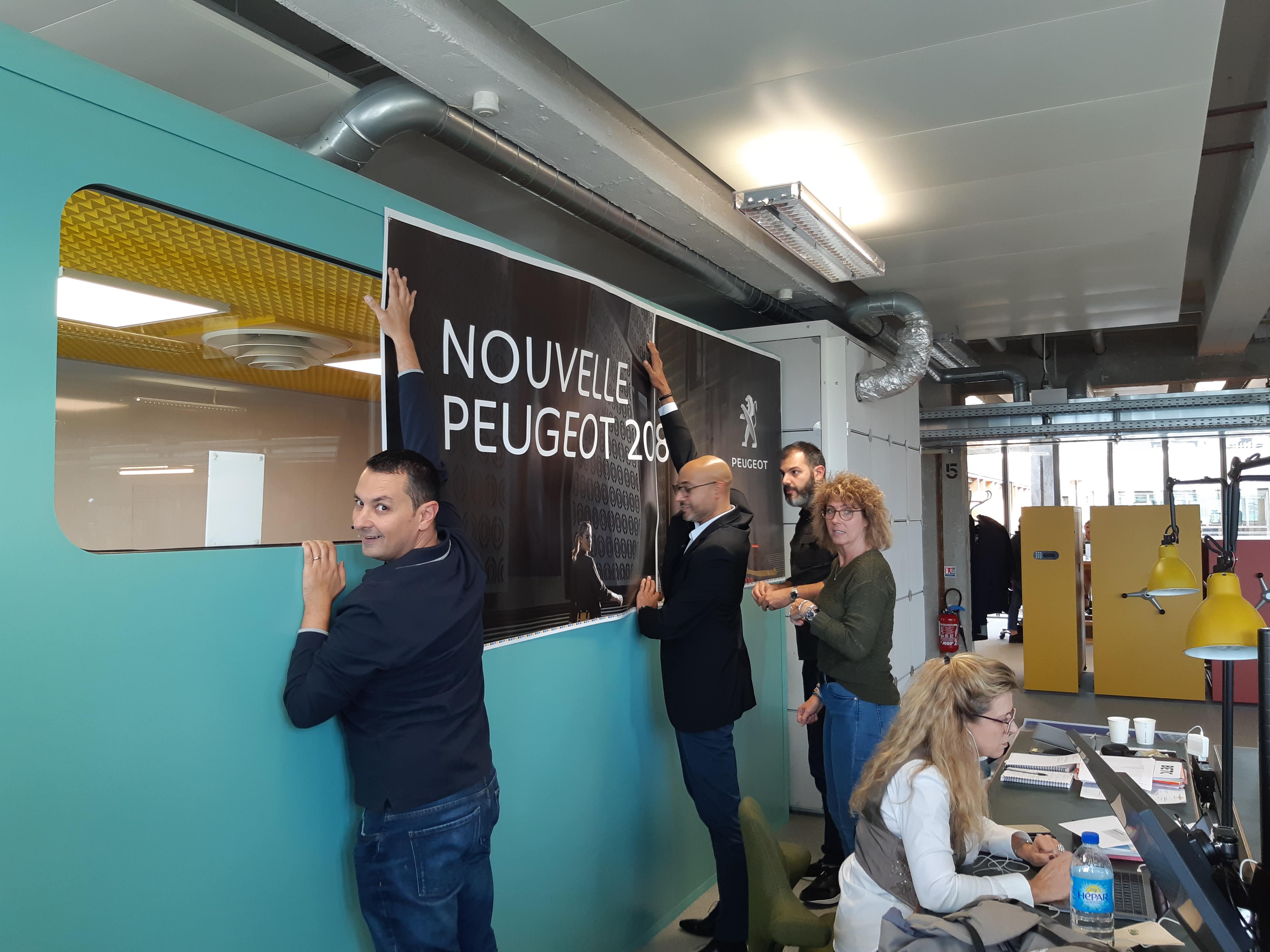 Employés installant une grande affiche “Nouvelle Peugeot 208” sur un mur de bureau moderne.