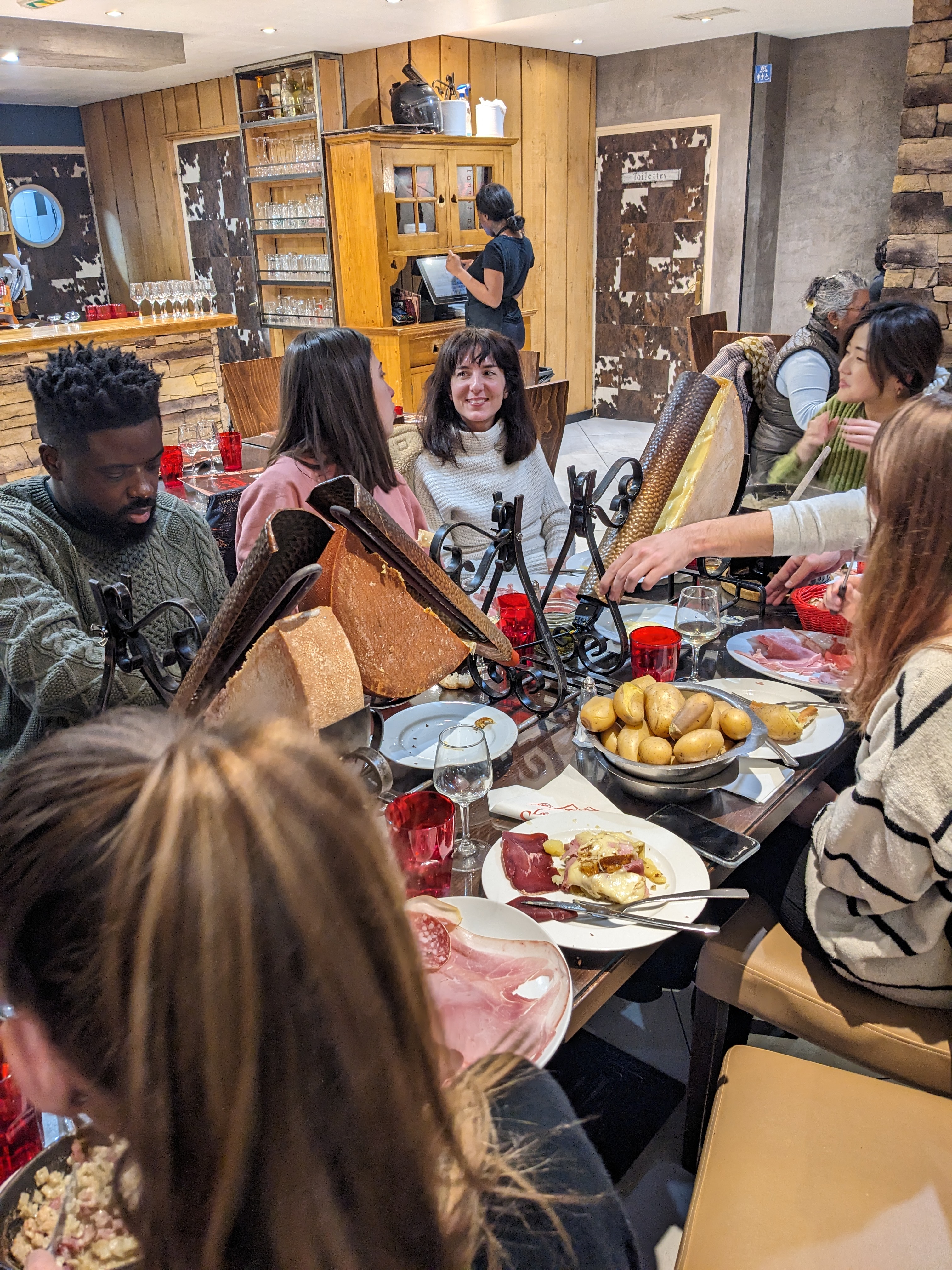Repas convivial autour d’un appareil à raclette, pommes de terre et charcuterie sur la table.