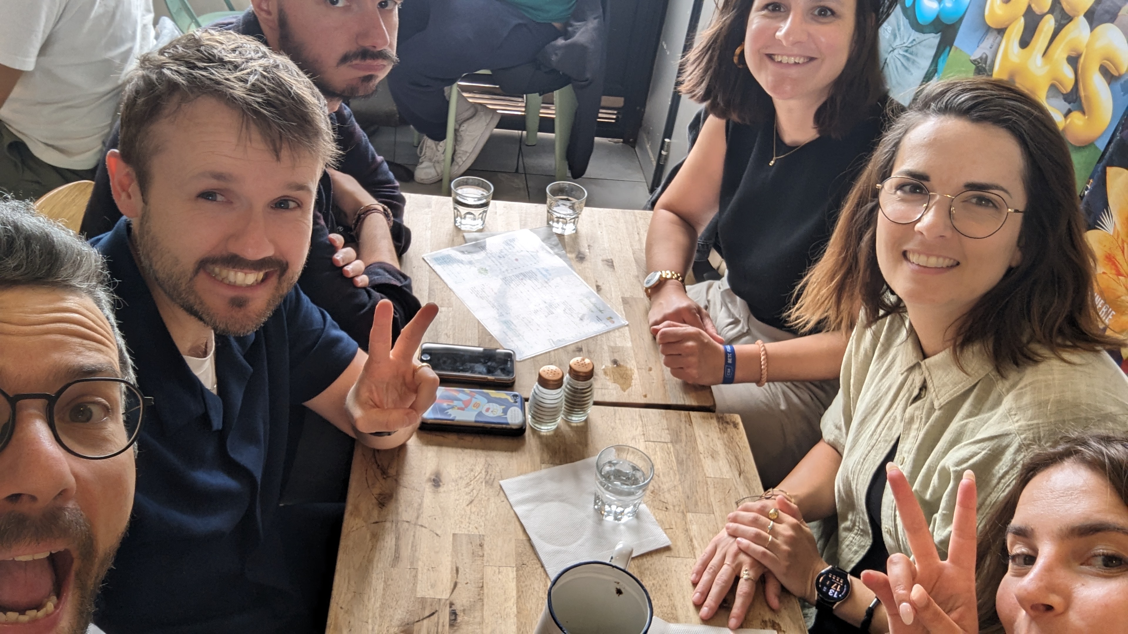 Selfie de groupe autour d’une table de café, plusieurs personnes souriantes faisant le signe de la paix.