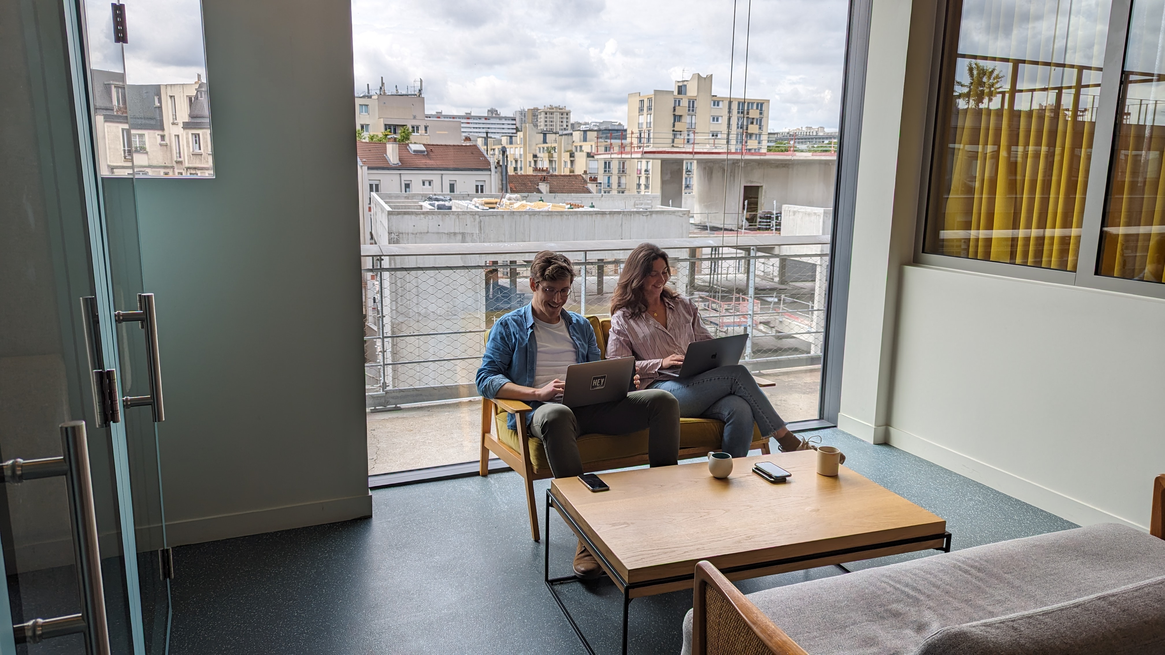 Deux collègues assis sur un canapé devant une baie vitrée, travaillant chacun sur un ordinateur portable.