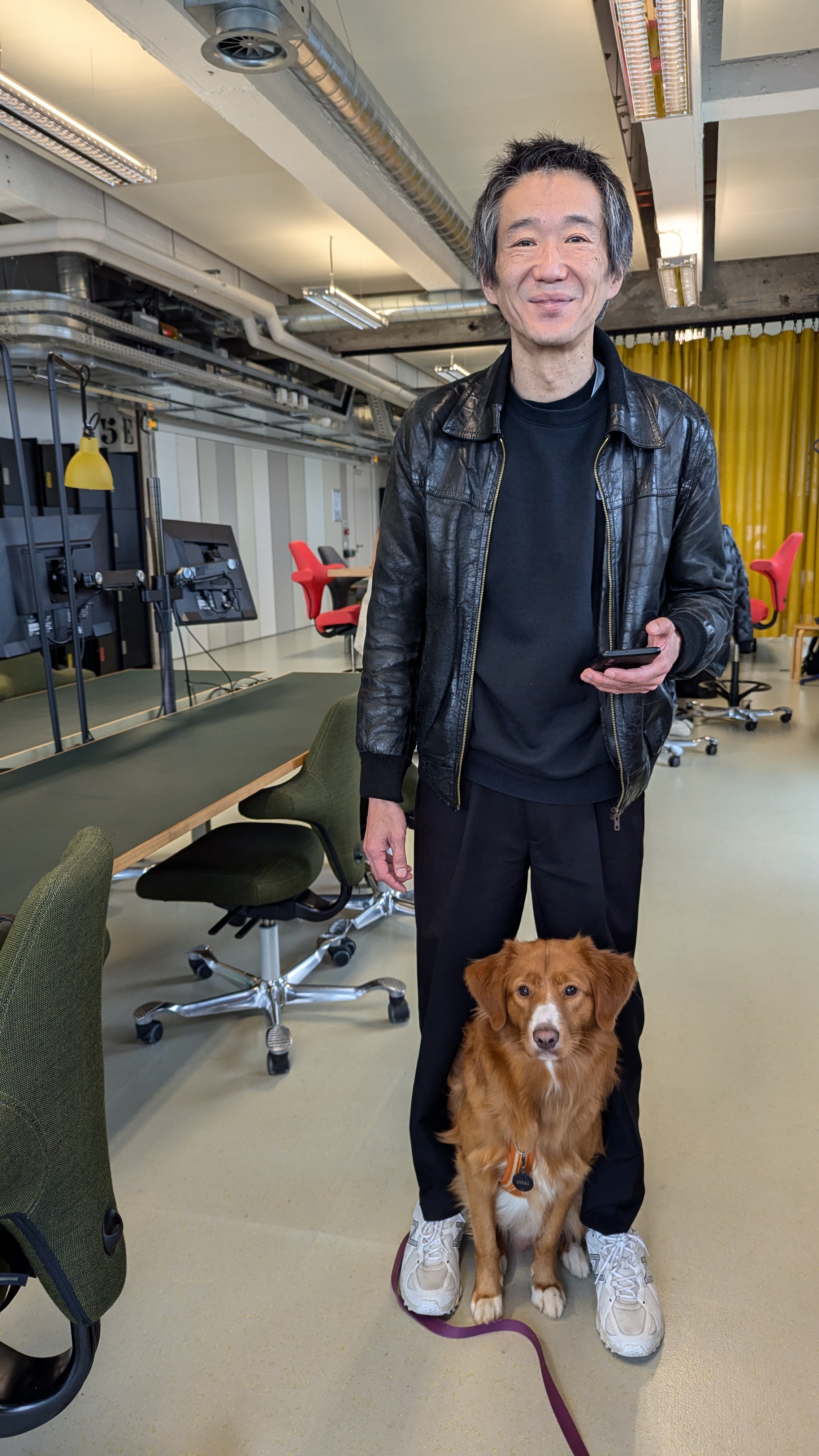 Portrait d’un homme debout dans un open space, tenant un téléphone, avec un chien roux assis à ses pieds.